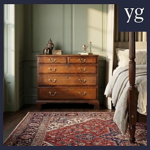 Puede incluir: Una cómoda de madera con cinco cajones y herrajes de latón se encuentra en un dormitorio. La cómoda es de madera de tonos cálidos y está coronada con objetos decorativos. Una alfombra estampada en rojo, azul y crema cubre el suelo. Se ve una cama con dosel.