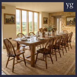 May include: A long rectangular wooden dining table with a natural finish, surrounded by ten matching wooden spindle-back chairs. The table is set with white plates, clear glassware, and decorative vases containing greenery and dried flowers.