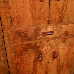 May include: Close-up of a wooden cabinet with a warm, reddish-brown finish. The cabinet features multiple doors and drawers with matching red handles. The wood grain is prominent, creating a textured appearance.