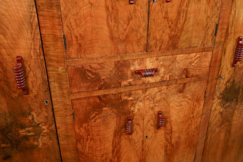 May include: Close-up of a wooden cabinet with a warm, reddish-brown finish. The cabinet features multiple doors and drawers with matching red handles. The wood grain is prominent, creating a textured appearance.