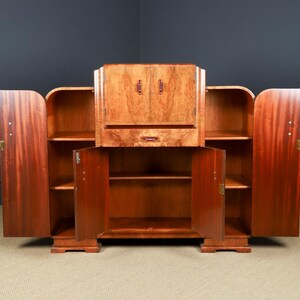 May include: A large, vintage wooden cabinet with multiple storage compartments. The cabinet features a central section with a drop-down desk and two side sections with open shelving. The doors and drawers have brass hardware. The wood has a rich, reddish-brown color.