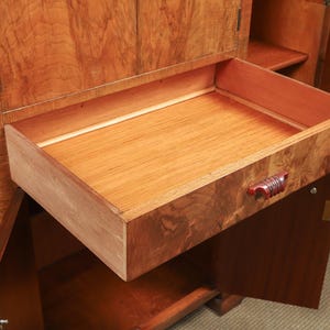 May include: A wooden cabinet with an open drawer. The drawer is made of light-colored wood with a reddish-brown handle. The cabinet has a rich, warm wood grain finish, and the drawer is partially extended.