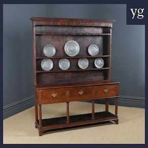 May include: A dark wood antique dresser with a top section for plates. The dresser has three drawers with brass handles and a lower shelf. The plates are round and silver. The dresser is a classic piece of furniture, a traditional design.