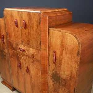 May include: A wooden cabinet with a warm, reddish-brown finish. The cabinet has a curved design with multiple doors and drawers. Red handles and pulls provide a contrasting accent. The wood grain is visible throughout the piece.