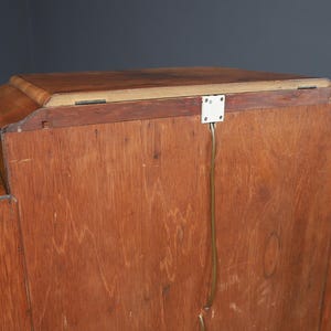 May include: A wooden cabinet with a light brown finish. The cabinet has a rectangular top and a series of vertical panels. A white electrical outlet is attached to the back with a wire running down. The cabinet is against a dark gray background.