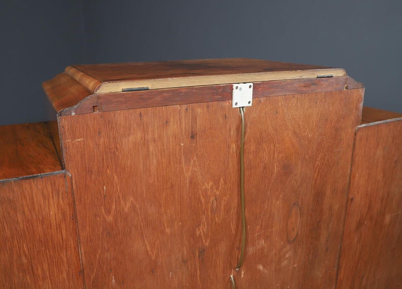 May include: A wooden cabinet with a light brown finish. The cabinet has a rectangular top and a series of vertical panels. A white electrical outlet is attached to the back with a wire running down. The cabinet is against a dark gray background.