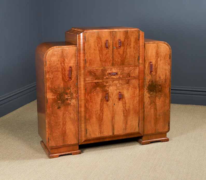 May include: A vintage wooden cabinet with a warm, reddish-brown finish. The cabinet features a central section with doors and a drawer, flanked by curved side panels. It has decorative hardware and a stepped top.
