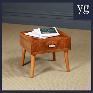 May include: A small wooden side table with a single drawer and four tapered legs. The table is made of a warm-toned wood with a visible grain pattern. A stack of magazines rests on the tabletop. The drawer has a silver handle.