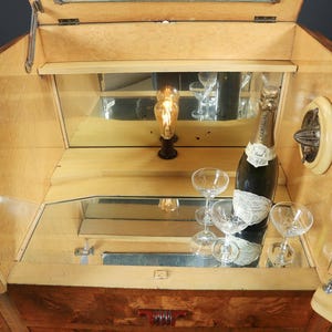 May include: Wooden Art Deco bar cabinet with mirrored interior. The cabinet is open, revealing a bottle of champagne, several champagne glasses, and a decorative light. The cabinet has a light wood finish and chrome hardware.
