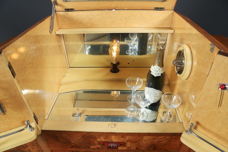 May include: Wooden Art Deco bar cabinet with mirrored interior. The cabinet is open, revealing a bottle of champagne, several champagne glasses, and a decorative light. The cabinet has a light wood finish and chrome hardware.
