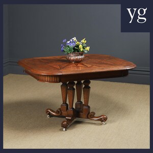May include: A dark wood table with a geometric inlay pattern on the top. The table has a pedestal base with four turned legs and four castors. A small copper planter with colorful flowers sits on the table.