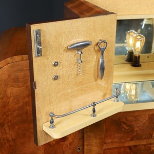 May include: A vintage wooden bar cabinet with a light-colored interior. The cabinet door is open, revealing a corkscrew, bottle opener, and a small shelf. The interior has a mirrored back and a glass.
