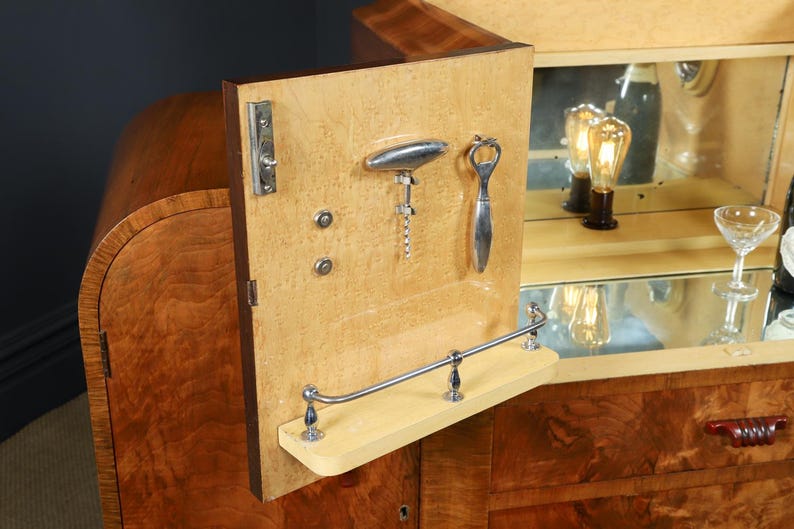May include: A vintage wooden bar cabinet with a light-colored interior. The cabinet door is open, revealing a corkscrew, bottle opener, and a small shelf. The interior has a mirrored back and a glass.