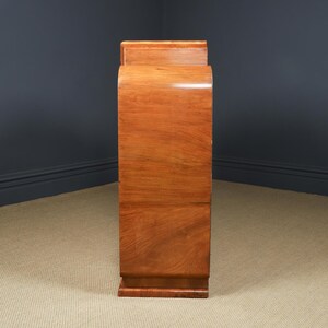 May include: A tall, wooden cabinet with a curved front and a rectangular top. The cabinet is made of a light brown wood with a glossy finish. The design is Art Deco style, with a streamlined, elegant appearance.