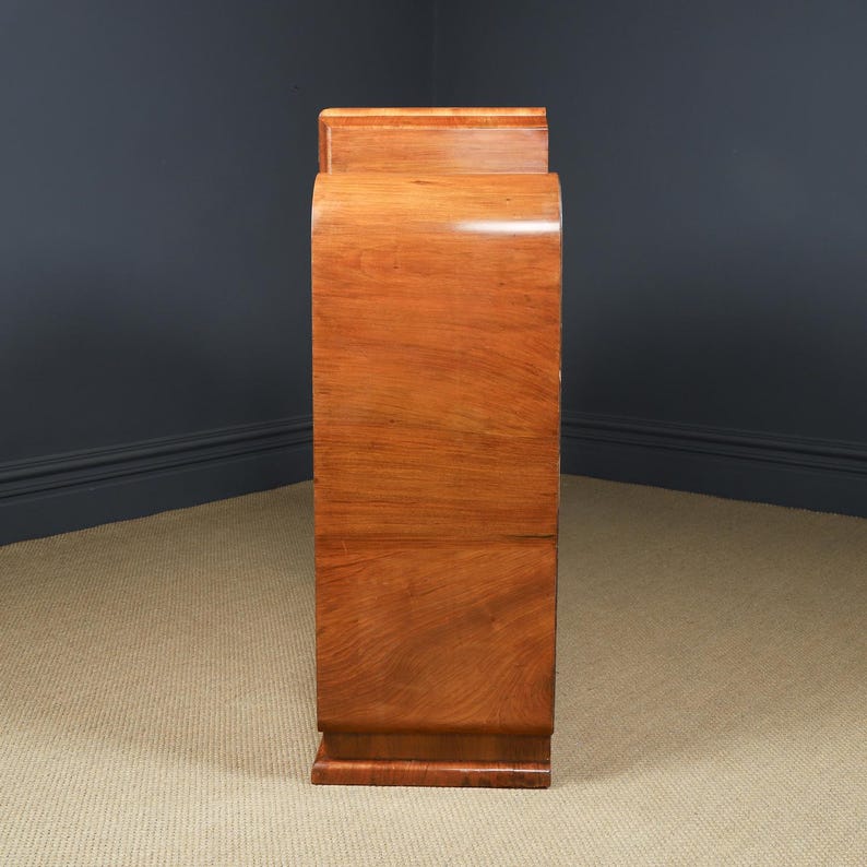 May include: A tall, wooden cabinet with a curved front and a rectangular top. The cabinet is made of a light brown wood with a glossy finish. The design is Art Deco style, with a streamlined, elegant appearance.