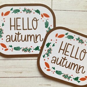 May include: Two brown rectangular signs with white backgrounds and a green and orange leaf border. The signs say "Hello autumn" in white text.