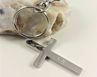 Puede incluir: Llavero de plata con un dije de cruz y la palabra "Jesús" grabada. El llavero tiene un anillo de plata y un clip de plata.