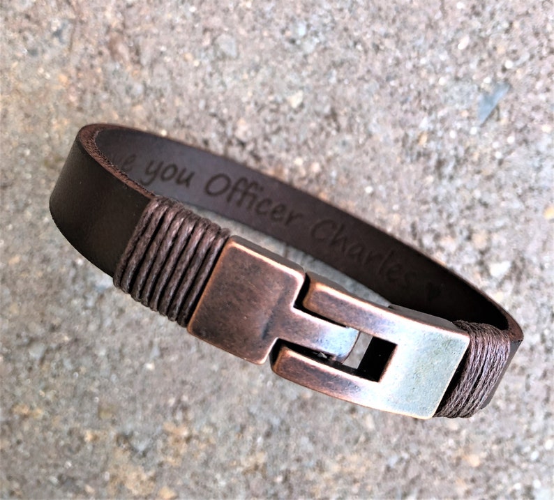 May include: Brown leather bracelet with a copper clasp and a braided cord detail. The bracelet is engraved with the text "I love you Officer Charles".
