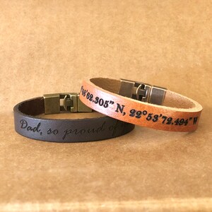 Può includere: Due braccialetti in pelle con testo inciso. Il braccialetto marrone dice "Papà, così orgoglioso di te". Il braccialetto beige dice "48°62.305" N, 22°53'72.494" W".