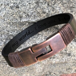 May include: A black leather bracelet with a copper clasp. The bracelet has a braided leather design and is engraved with the words "You are my world... I love you."