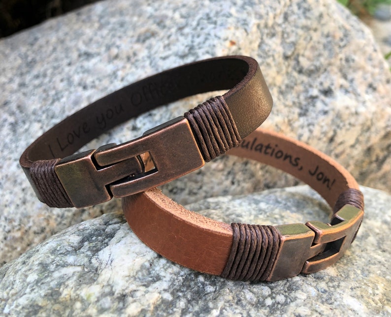 May include: Two brown leather bracelets with metal clasps. One bracelet is engraved with "I love you Offi", and the other is engraved with "Congratulations, Jon!"