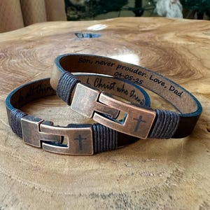 May include: Two dark brown leather bracelets featuring copper-toned metal clasps with an engraved cross symbol. The interior of the leather bands is inscribed with the text: Son, never prouder. Love, Dad 04-05-25 and do all things through Christ who strengthens.