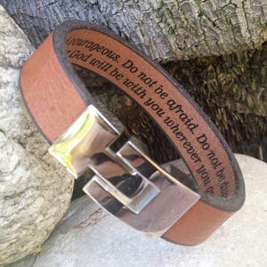 Puede incluir: Pulsera de cuero marrón con cierre de plata. La pulsera está grabada con el texto "No tengas miedo. No te desanimes. Dios estará contigo dondequiera que vayas."