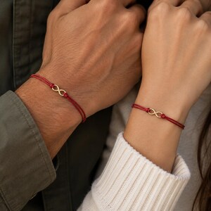 May include: Two matching bracelets featuring a thin red cord and a small gold-toned infinity symbol charm. Each bracelet is secured with a simple knot design, showcasing a minimalist aesthetic with a vibrant red color and a metallic gold accent.