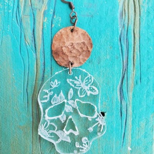 Może przedstawiać: Para kolczyków wiszących z przezroczystą czaszką z akrylu z kwiatowymi detalami i brązowym metalowym dyskiem.