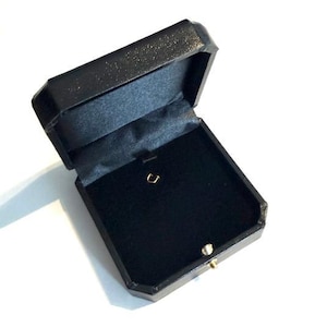May include: A black jewelry box with an octagonal shape and a hinged lid. The interior is lined with black velvet and features a small gold-colored metal hook for securing a piece of jewelry. The exterior has a textured finish and a gold latch.