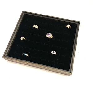 May include: A wooden ring display case with a black velvet interior. The case holds several rings of various designs, including gold bands with gemstones. The rings are arranged in individual slots, showcasing jewelry.