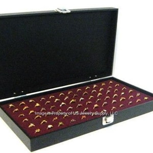 May include: Black ring display box with a burgundy velvet interior. The box has a hinged lid and a lock. The box is filled with empty ring slots.