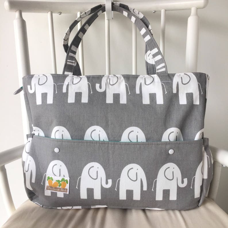 May include: A gray tote bag with a white elephant pattern. The bag has a zipper closure and a large pocket on the front. The bag has a white label with the text "The Crafty Stitch" on the front.