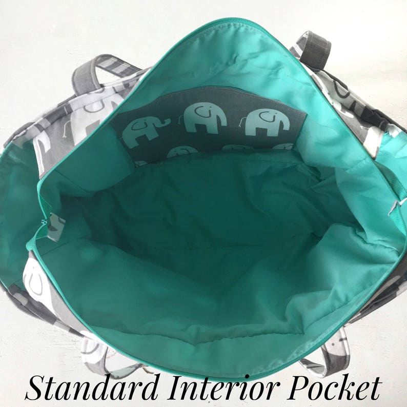 May include: The inside of a gray and white tote bag with a teal lining. The bag has a zippered pocket with a gray background and a pattern of white elephants. The pocket is labeled "Standard Interior Pocket".