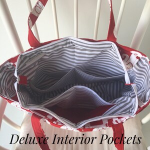 May include: Red and white tote bag with a striped gray and white lining. The bag has multiple interior pockets. The text "Deluxe Interior Pockets" is printed below the bag.