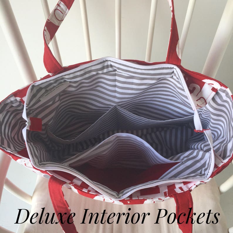May include: Red and white tote bag with a striped gray and white lining. The bag has multiple interior pockets. The text "Deluxe Interior Pockets" is printed below the bag.