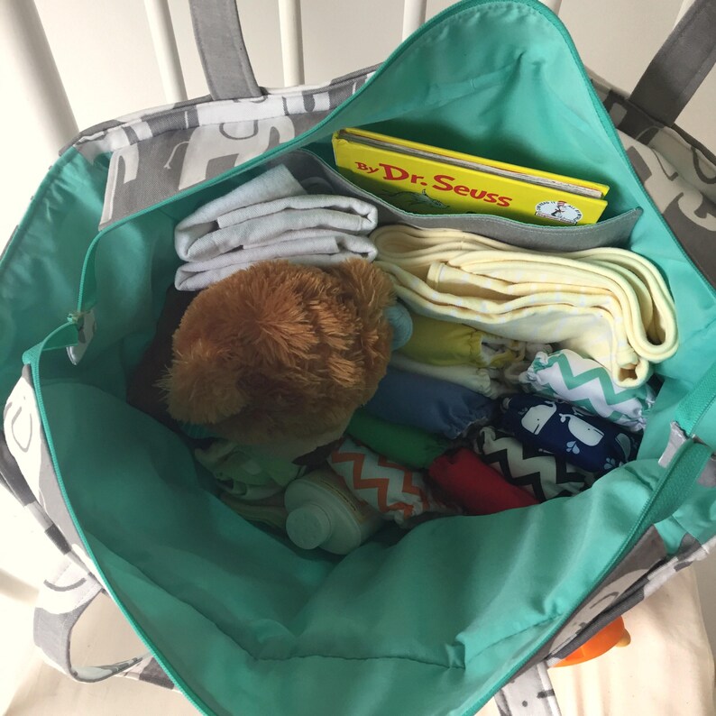 May include: A turquoise and gray tote bag with a zipper closure and handles. The bag is filled with various items, including a stuffed animal, a book titled "By Dr. Seuss", and several folded cloths.