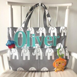 May include: Gray and white diaper bag with a personalized name "Oliver" in teal letters. The bag has multiple pockets and features a pattern of white elephants on a gray background. A toy is attached to the bag.