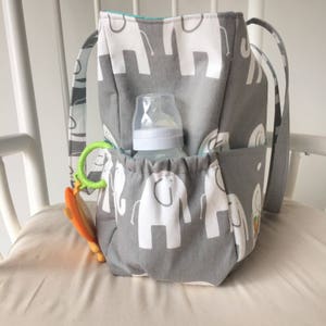 May include: A gray and white diaper bag with elephant print. The bag has multiple pockets and a bottle holder. A baby bottle and a green and orange toy are in the bag.