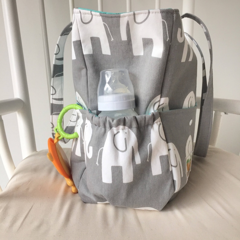 May include: A gray and white diaper bag with elephant print. The bag has multiple pockets and a bottle holder. A baby bottle and a green and orange toy are in the bag.
