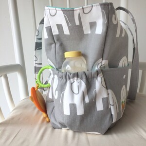 May include: A gray and white diaper bag with a pattern of elephants. The bag has multiple pockets and a drawstring closure. A clear plastic bottle and a green and orange toy are inside the bag.