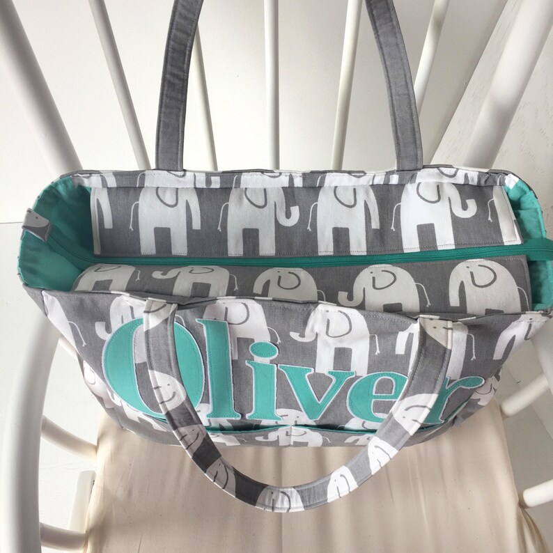 May include: Gray and teal tote bag with a zippered top and white elephant print. The bag has the name "Oliver" embroidered on the front in teal.