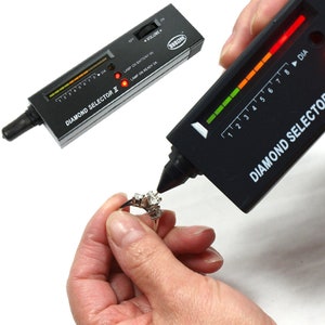 May include: A black diamond tester with a green and red LED display. The tester is being used to test a ring with a diamond.
