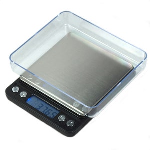 May include: A black digital scale with a stainless steel weighing surface and a clear plastic cover. The display shows a weight of 37.65. The scale has several buttons and is designed for precision measurements.