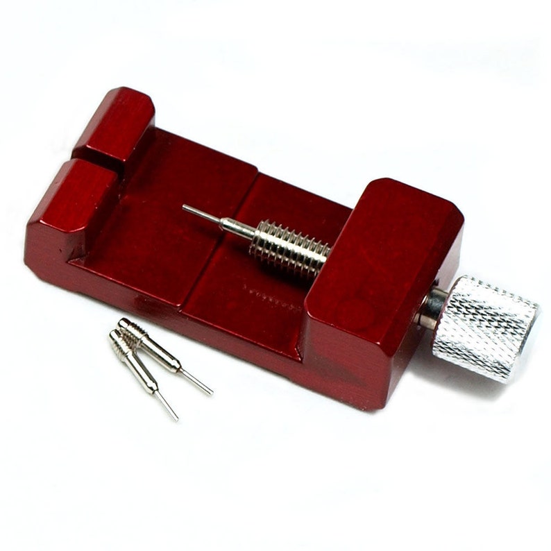 May include: A red wooden watch band link remover tool with a silver metal screw and two small silver pins. The tool is designed to remove links from watch bands. The wooden base is rectangular with a slot for the watch band.