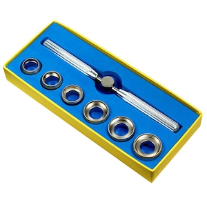May include: A silver watch case opener tool with a blue and yellow case. The tool has a handle with a grooved design and a metal head. The case includes six different sized rings for opening watch cases.