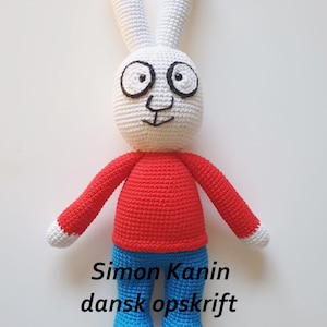May include: A crocheted white rabbit with large black eyes and a red shirt and blue pants. The rabbit is standing on a white background. The text "Simon Kanin dansk opskrift @ti_tommelfingre" is written below the rabbit.