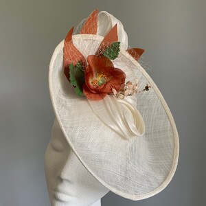 May include: A white fascinator hat with a large, orange flower and green leaves. The hat is made of a delicate, woven material and has a wide brim.