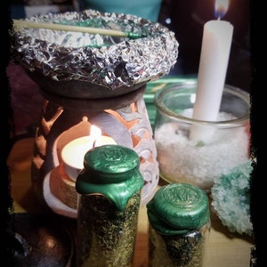 Op de afbeelding: Twee kleine glazen potten met groene waszegels, gevuld met gedroogde kruiden, staan op een houten oppervlak. Op de achtergrond staan een witte kaars en een kleine, ronde, keramische wierookbrander.