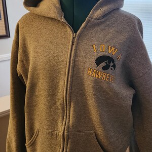 May include: A gray zip-up hoodie with a hood, front pockets, and the words "IOWA HAWKEYES" printed on the front. The hoodie is displayed on a mannequin. The zipper is gold-colored.
